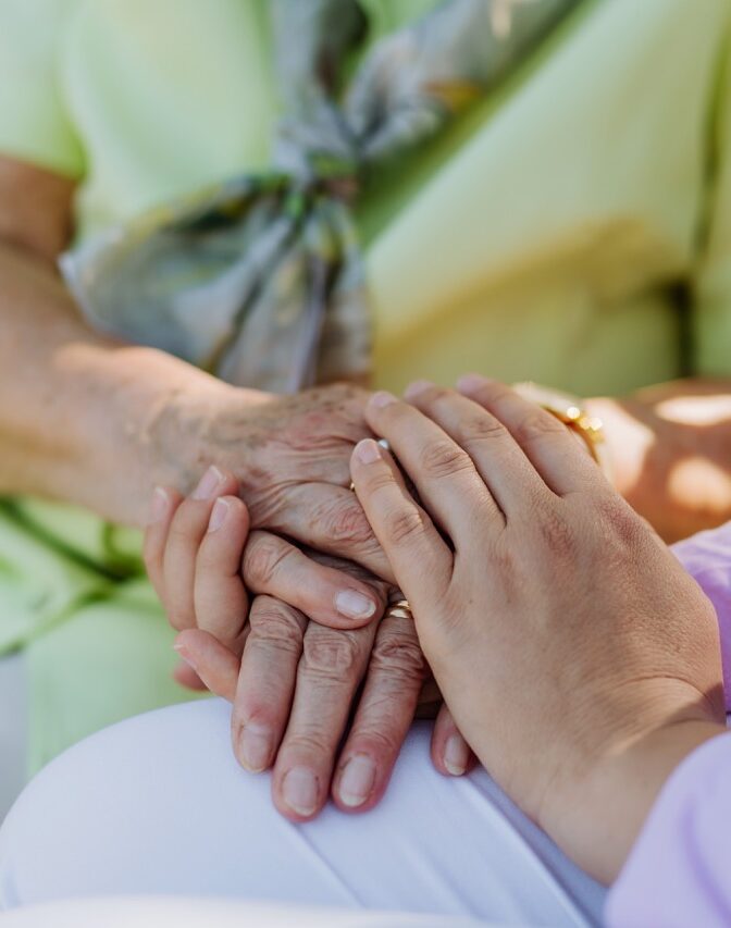 A close-up of caregiver consoling senior woman and touching her hand when sitting on bench in park in summer.